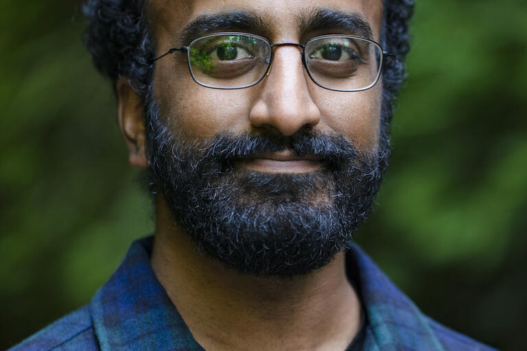 Headshot of Aanan Varma. He is outside and smiling. He has short curly black hair, a short black beard, and a medium brown skin tone. He is wearing eyeglasses and a blue and green plaid button-up collared shirt. The background is out of focus.