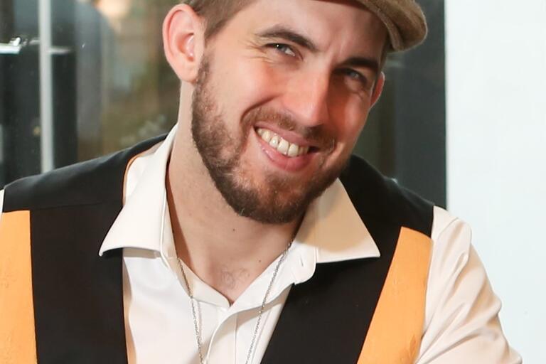 Headshot of Brian Sonia-Wallace. He is indoors and smiling. He has a short brown beard, short brown hair, and a pale skin tone. He is wearing a brown newsboy hat, a yellow vest, and a white collared shirt. He is sitting at a desk.