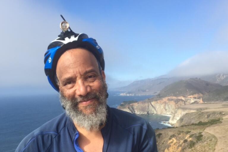Selfie of C.S. Giscombe, a Black American poet. He is outdoors and smiling. He has a long salt-and-pepper beard and a light brown skin tone. He is wearing a blue helmet and blue bike-riding shirt. The scenery in the background is beautiful.