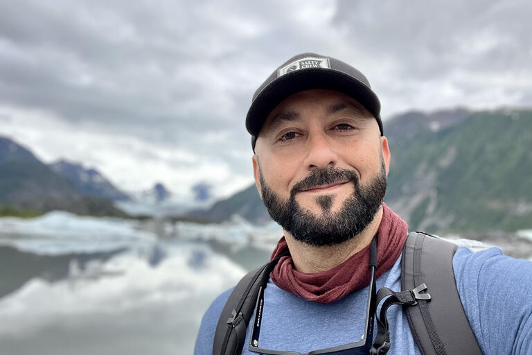Selfie of Craig Santos Perez, a Pacific Islander from Guam. He is outdoors and smiling. He has a short black beard and a light almond skin tone. He is wearing a black baseball hat, brick red neck scarf, denim blue t-shirt, and black backpack.