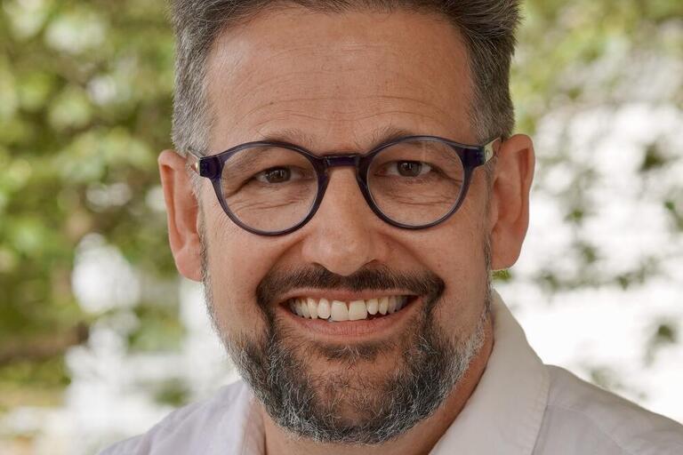Headshot of David Hassler. He is outdoors and smiling. He has a short salt-and-pepper beard, short gray hair, and a light, tanned skin tone. He is wearing eyeglasses and a white collared shirt. He is under the canopy of green trees.
