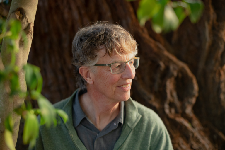 Candid photo of John Shoptaw. He is outdoors and smiling. He has short gray hair and a rosy pale skin tone. He is wearing eyeglasses, a gray polo shirt, and a sage green sweater. He is standing close to a redwood tree and looking away.