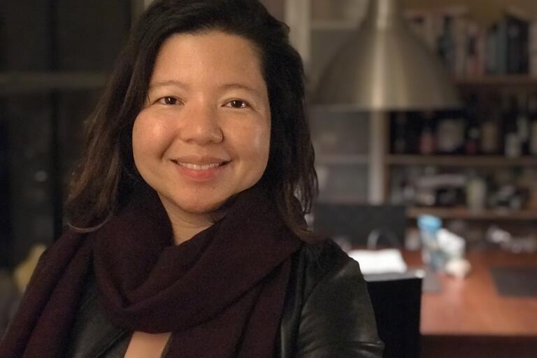 Selfie of Karen Llagas, a Filipino and American poet. She is indoors and smiling. She has straight, long dark hair and a light almond skin tone. She is wearing a dark wine red scarf and black leather-like jacket. The background is out of focus.