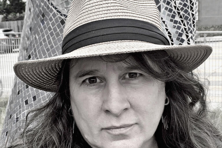 Selfie of Kim Shuck, a Cherokee Nation poet. She is outdoors and looking into the camera seriously. She is wearing a straw hat, heather sweater, and black collarless blouse. Photo is almost black and white with a slight blue tint.