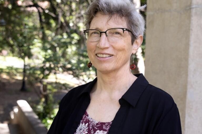 Headshot of Linda Rugg, a Swedish American scholar. She is outside and smiling. She has short gray hair and a pale skin tone. She is wearing eyeglasses, a black collared shirt unbuttoned over a burgundy floral blouse. The background is out of focus.