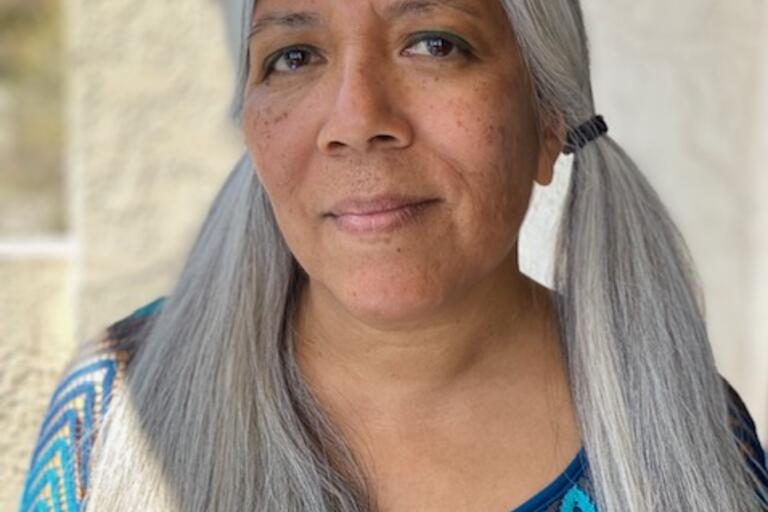 Headshot of Maw Shein Win, a Burmese American poet. She is outside and smiling. She has long, straight white hair in pigtails and a light brown skin tone. She is wearing a blue collarless blouse with a chevron pattern. The background is out of focus.