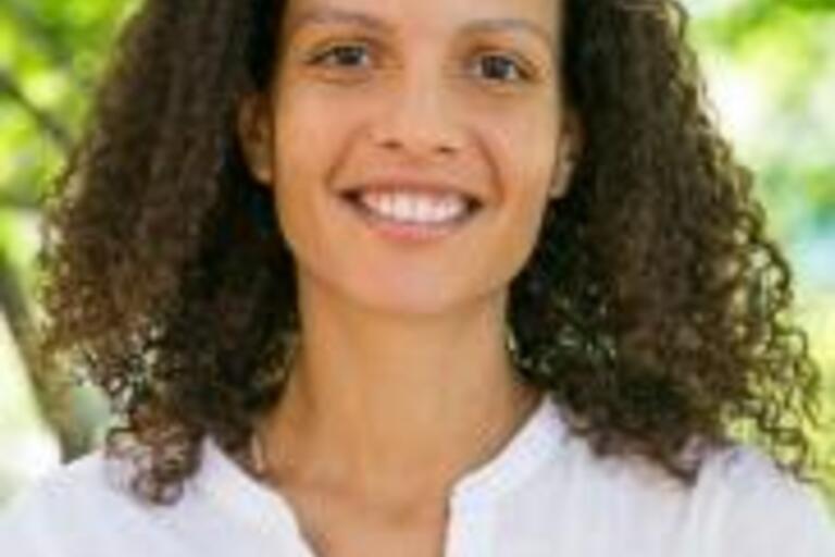 Headshot of Sarah Vaughn. She is outside and smiling. She has long curly brown hair and a light brown skin tone. She is wearing a collarless white blouse. She is under the canopy of green trees and the background is out of focus.