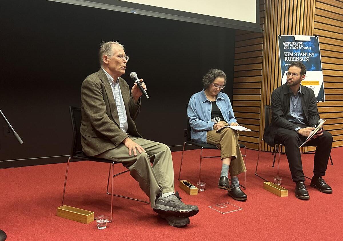 Photo of 3 people sitting on a stage with a black background and red carpet. Photo is close up but shows all seated figures well. Left to right are: Kim Stanley Robinson, Katherine Snyder, and Daniel Aldana Cohen.