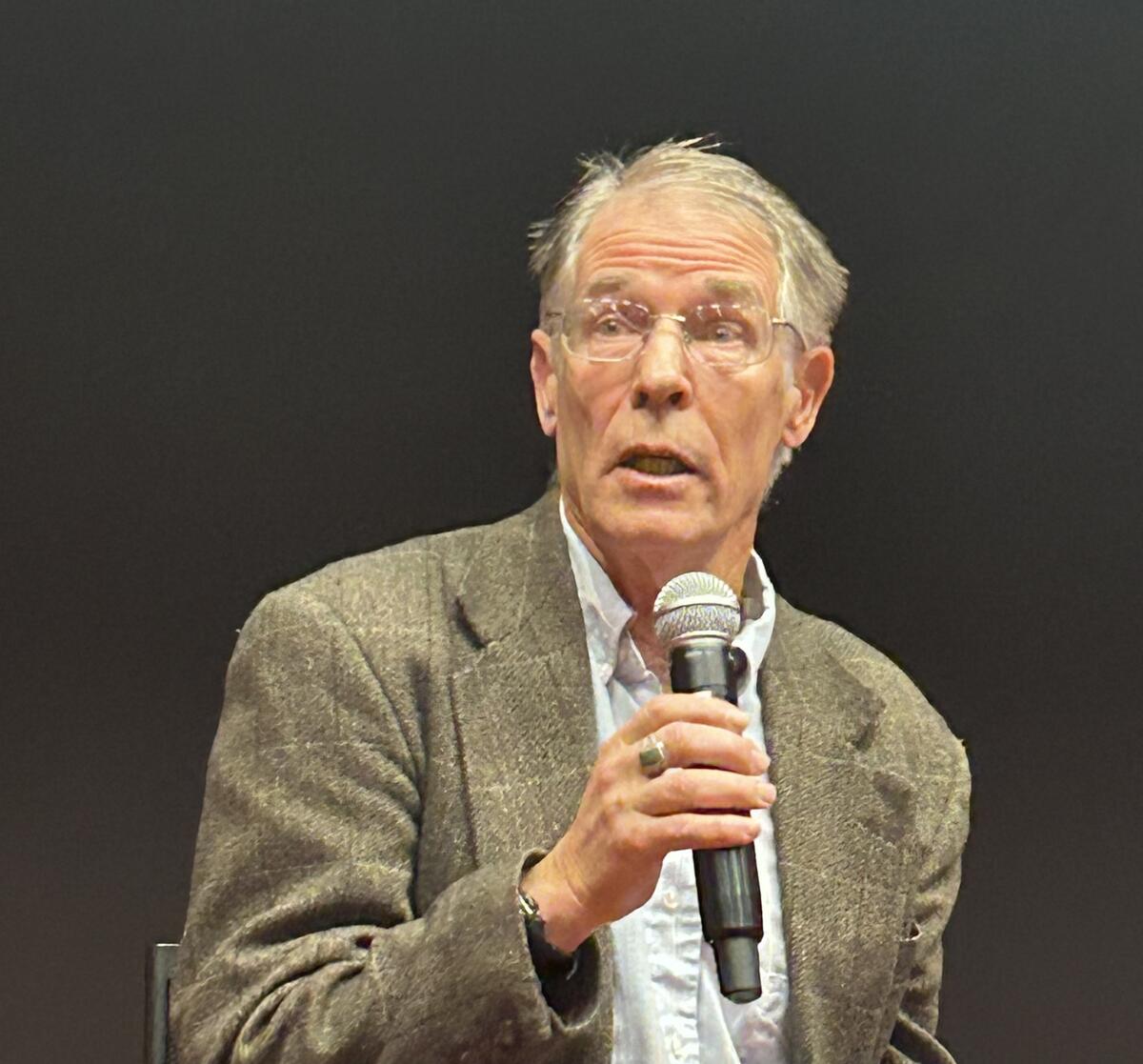 Photo of Kim Stanley Robinson, a man with a pale skin tone and gray hair, speaking on stage and holding a microphone. He is wearing a light gray blazer and a light blue collar shirt. He wears glasses.