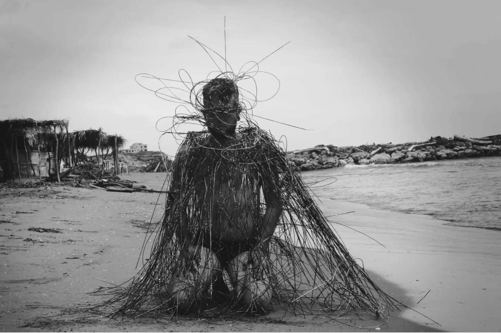 Black and white image of a man kneeling in the sand on a beach. He is covered head to toe by cables draped over his body. 